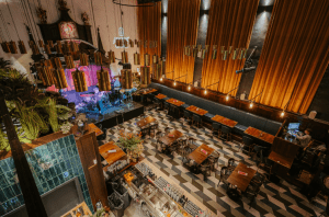 Interior view of Amazing Grace London, featuring a stylish stage setup with musical instruments, warm lighting, large gold curtains, modern hanging light fixtures, patterned tile flooring, and neatly arranged wooden tables and chairs in a lively, contemporary venue.