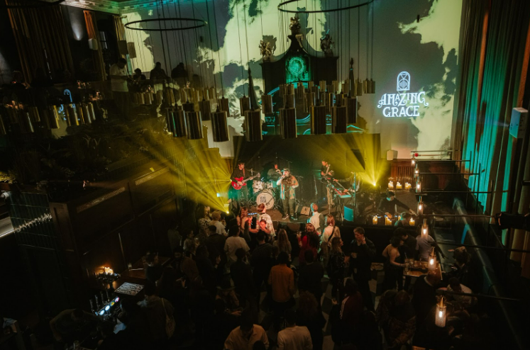 Live music performance at Amazing Grace London, showing a lively crowd gathered around a stage illuminated by colorful lights, with musicians playing under large hanging gold fixtures and the venue’s name projected on the wall.